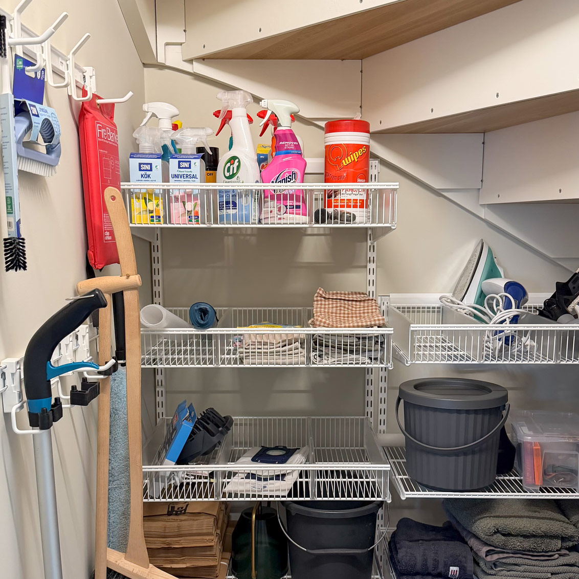 Elfa hyllor under en trappa med städprodukter.