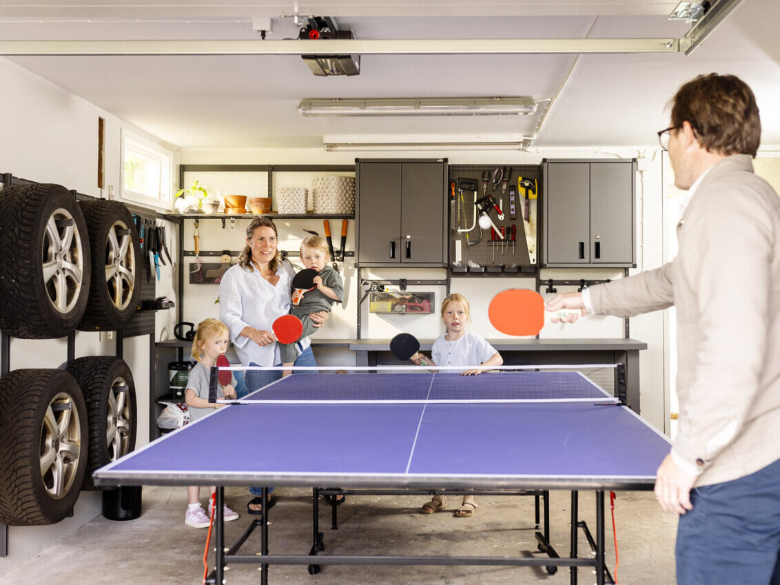 The family plays table tennis in the middle of the garage around a table tennis table in front of Garage+ storage from Elfa.