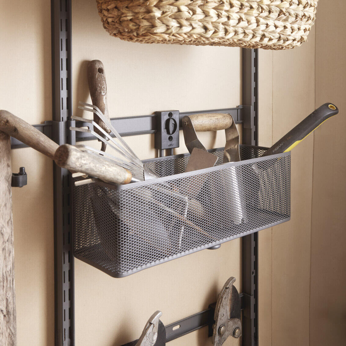 Garage+ storage track from Elfa, where a mesh basket hangs with gardening tools.
