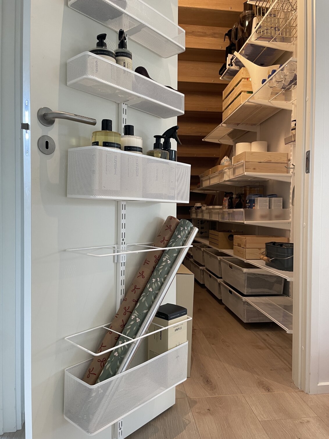 Förråd under en trappa med förvaring från Elfa på väggen och på dörren där det förvaras torrvaror, städsaker och presentpapper.