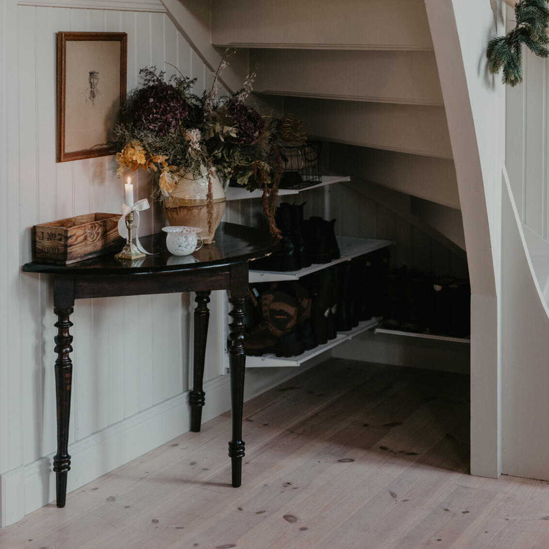 Space under a staircase with shoe shelves from Classic by Elfa in white and a table with decorations on it.