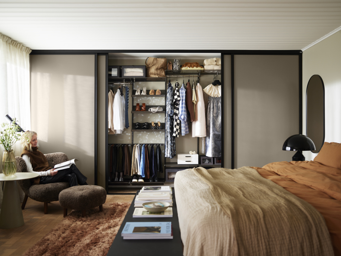 Kobieta siedzi na fotelu i czyta w sypialni z szarą szafą Classic by Elfa z brązowymi drzwiami przesuwnymi Artic.