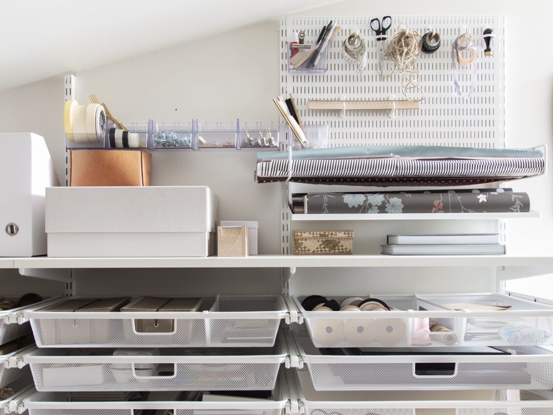 Viistokattoinen askarteluhuone, jossa on Classic by Elfa -säilytyskalusteita. Askartelutarvikkeet säilytetään koreissa ja säilytystaululla.