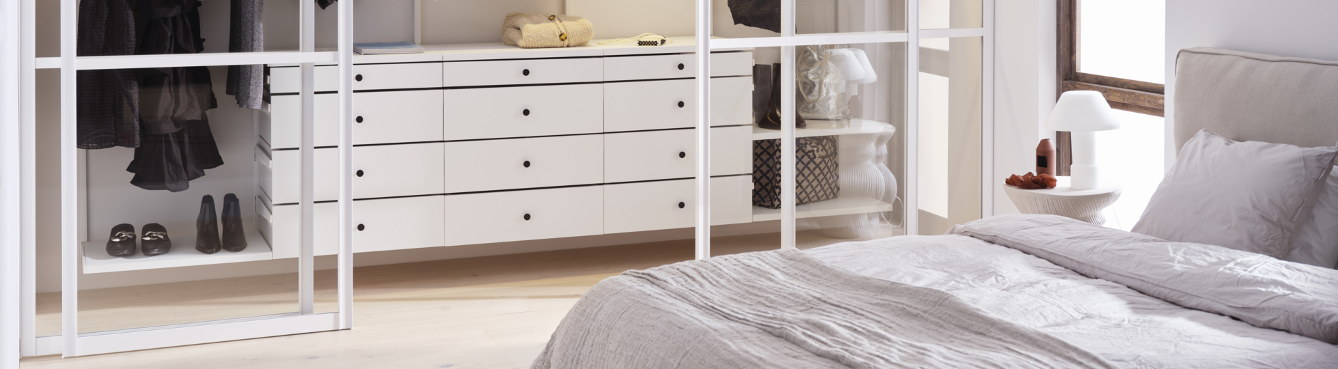 Sypialnia z białą szafą Décor+ by Elfa z wysuwanymi szufladami i szklanymi drzwiami przesuwnymi.