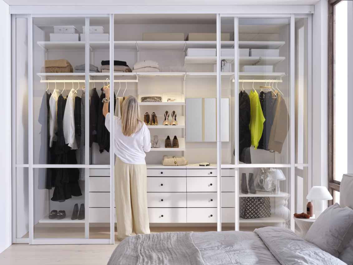 Ein Schlafzimmer, in dem sich eine Frau nach einem Pullover in einem weißen Décor+ by Elfa Kleiderschrank mit Schiebetüren mit Glasfüllung streckt.