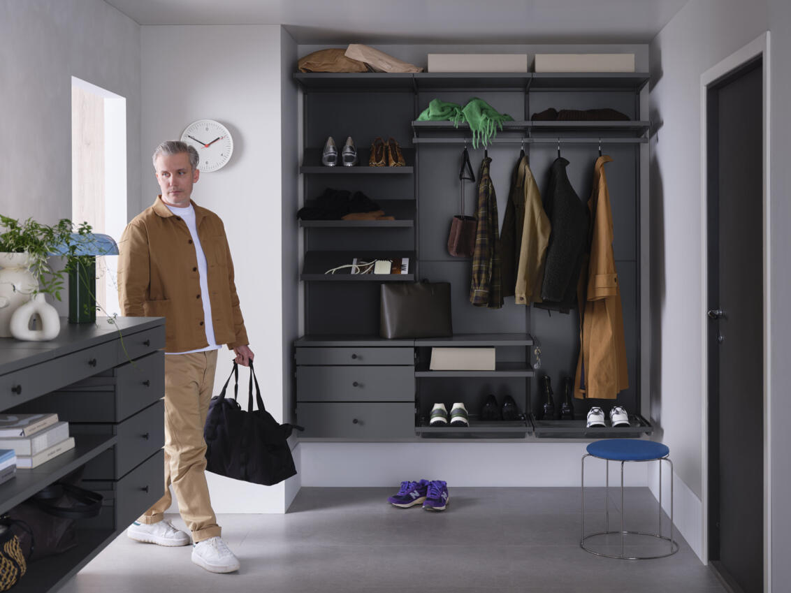 Mies astuu eteiseen, jossa on Elfan Décor+ vaatekaappi, jossa on ripustettuja vaatteita sekä harmaita viistoja hyllyjä, laatikoita ja kenkähyllyjä.