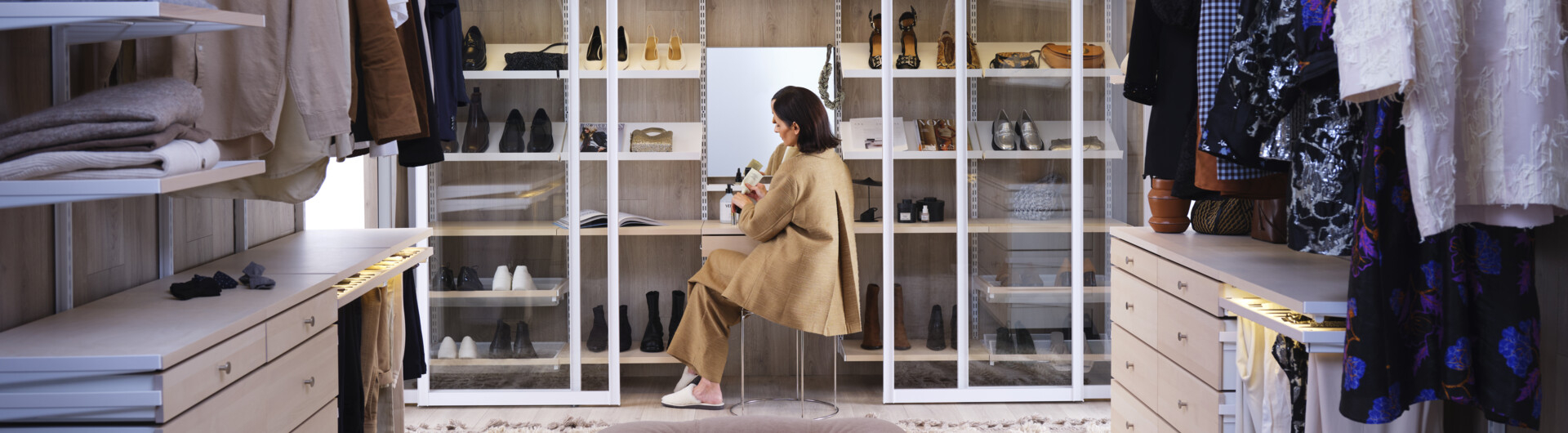 A woman is sitting in a walk-in closet with storage from Décor+ by Elfa in white and birch with glass sliding doors with white frames.