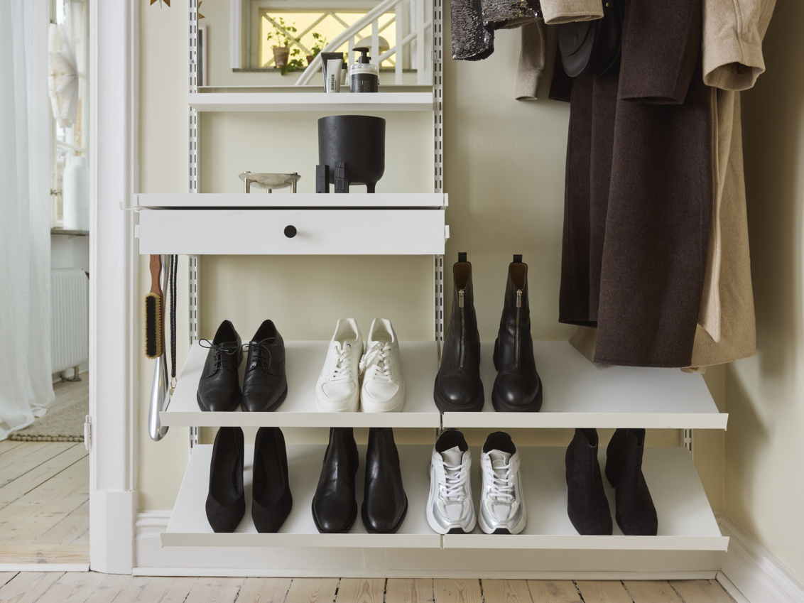 Pieni eteinen, jossa on valkoinen Décor+ by Elfa -vaatekaappi, jossa roikkuvat vaatteet ja kaltevat hyllyt kengät jalassa.