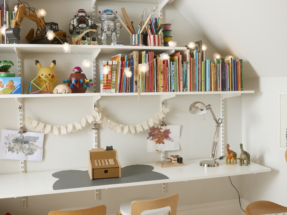 Pokój dziecięcy z półką na książki pod skośnym sufitem wypełnioną książkami i zabawkami. Dolna półka pełna stanowi biurko.