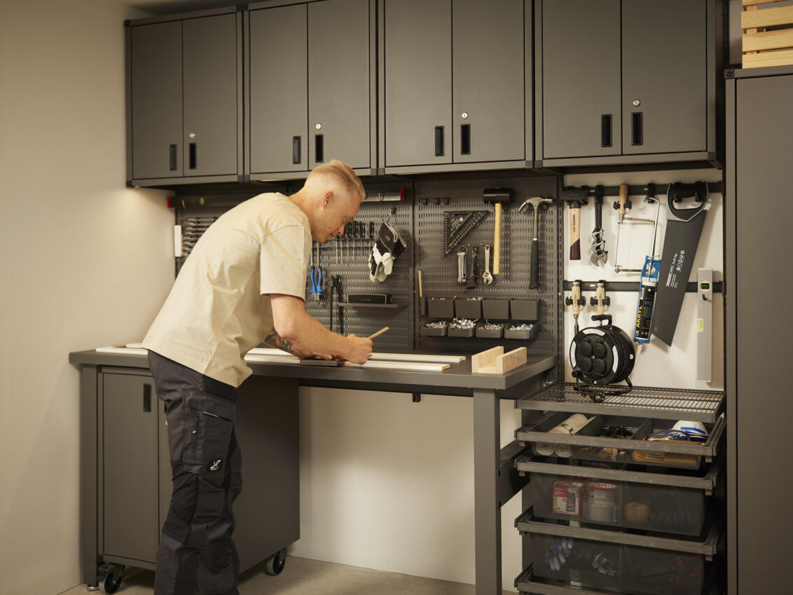 Un hombre trabaja en un banco de trabajo en una unidad de almacenamiento Garage+ de Elfa con armarios, estantes de almacenamiento y banco de trabajo.